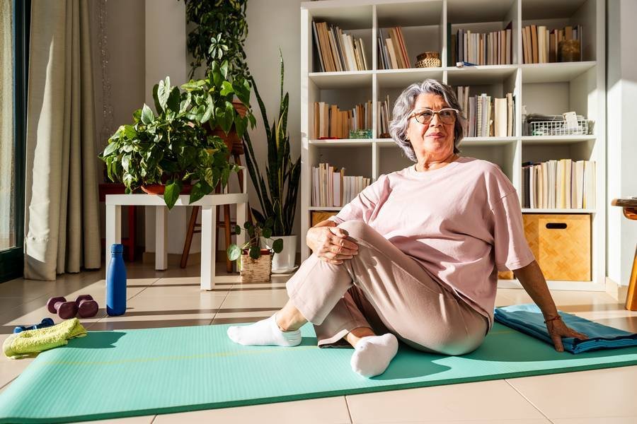 Senior woman sitting on yoga mat at home after stretching workout — fitness after 40