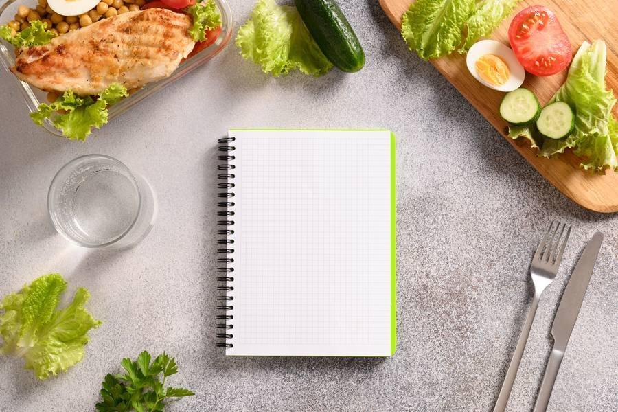 Healthy meal planning notebook with grilled chicken, vegetables, and eggs on kitchen counter